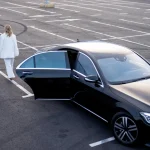 High angle view of a professional man in a black suit rolling a red suitcase alongside a woman in a white pantsuit as they walk away from a black luxury executive sedan with an open rear door in a parking lot